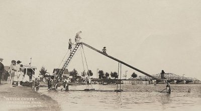 vintage photo of kids on a large water slide at "Wander Park" at the city's lake, with people standing around watching, in the style of late Victorian era cabinet card, early American photography, vintage typography that says "Water Chute", kids playing and laughing on the 50 foot tall slide, a bridge in the background.