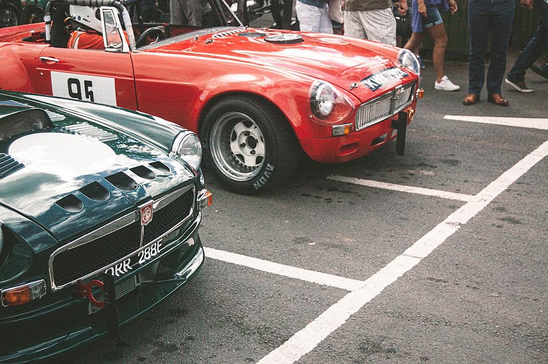 Close up of two classic cars in line at an event, a red MActionBar and green TVR Lightning, people around them, white lines on the asphalt floor, taken with a Canon EOS style camera in the style of Canon EOS.