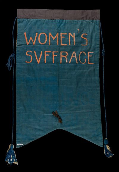 a banner with the words "WOMEN'Sglyph suff bureaucratsce" embroidered on it, blue fabric with dark tassels at each corner