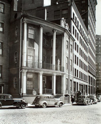 A vintage black and white photograph of a New York building facade with columns, architectural details, a brick wall, a stone balcony, in the classical architecture style, cars parked on the street in front, depicting bustling city life, in the style of architectural photography, in the style of architectural visualization, with highly detailed.