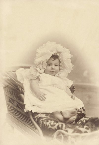 A baby in white dress and bonnet sitting on cradle, vintage photography