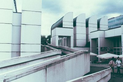 A grainy photo of the modernist museum at the art center in Tokyo, in the style of brutalism, with white concrete walls and curved structures, captured with an expired fujifilm for high contrast and muted colors, showing people walking around.