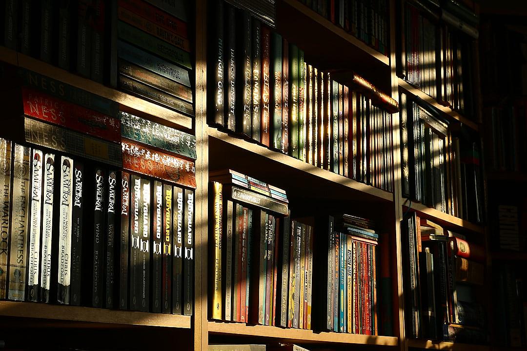 A closeup of bookshelves filled with various books, illuminated by the soft glow from sunlight streaming through a window. The books range in color and size, creating an array of textures on each shelf. In the background is darkness or shadows that suggest more shelves behind them. This scene captures a moment where you can feel your imagination burning as if looking into endless possibilities, highlighting how a single detail like light affects the overall mood and atmosphere.