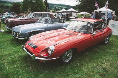 A photo of classic cars at an English car show in the countryside, including one red Jaguar E-type and two silver modern BMWs. The Jaguar E-type was photographed in the style of a classic British sports car, while the BMWs were captured in a more modern automotive design.