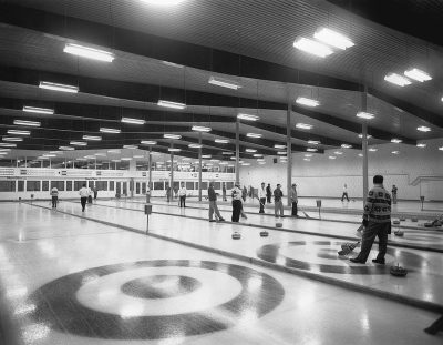 A wideangle black and white photo shows an indoor curling rink with people playing in the background. There is one large center circle where rocks are placed while standing at opposite ends. The bright lights are above them and the carpeted floor surrounds their paths. An overhead skylight illuminates everything. It looks like something from 'The Valentino Show' or the movie 'Onion motion picture'. It feels nostalgic and retro. Shot in the style of [Ansel Adams](https://goo.gl/search?artist%20Ansel%20Adams).