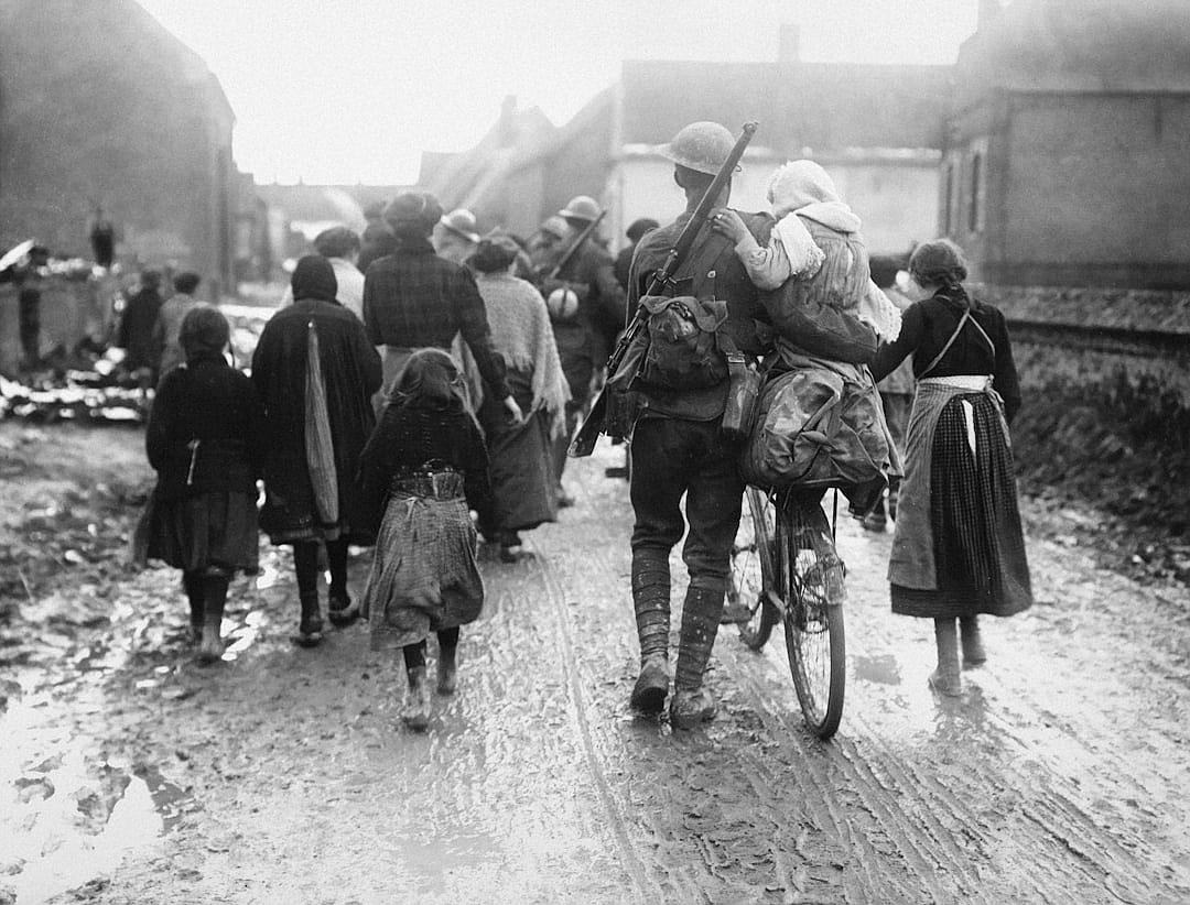 A soldier on his bike carries an armful of children, women and men walk behind him along the road in French village after battle, World War I photography, black & white photo