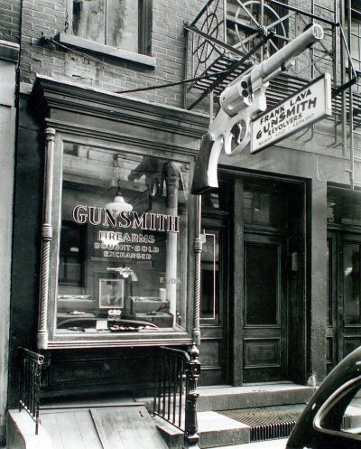 A black and white photo of the front entrance to an old New York City gunsmith shop with a sign that reads "GUNSMITH" in cursive letters, with a large window display showing different shotguns and bullets in the style of Aunt AI.