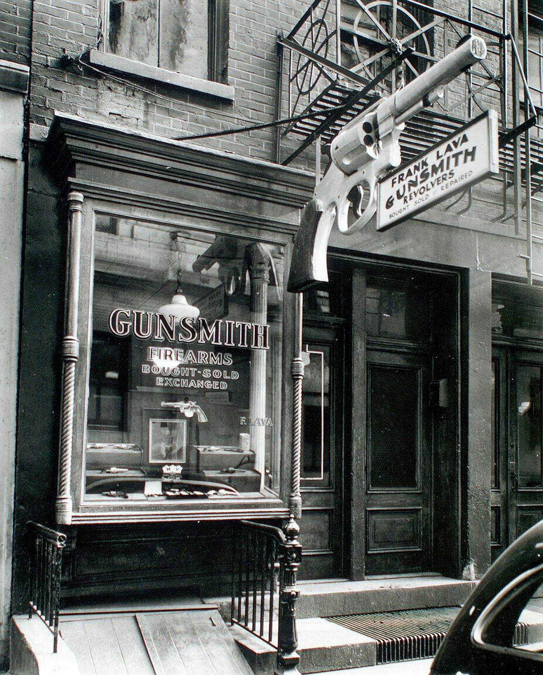 A black and white photo of the front entrance to an old New York City gunsmith shop with a sign that reads “GUNSMITH” in cursive letters, with a large window display showing different shotguns and bullets in the style of Aunt AI.