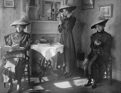 A black and white photograph shows three women in Edwardian , two sitting at a table with one standing between them holding an old-style telephone to her ear. They are inside the drawing room of their large house. The women have fur coats, hats, and gloves on.