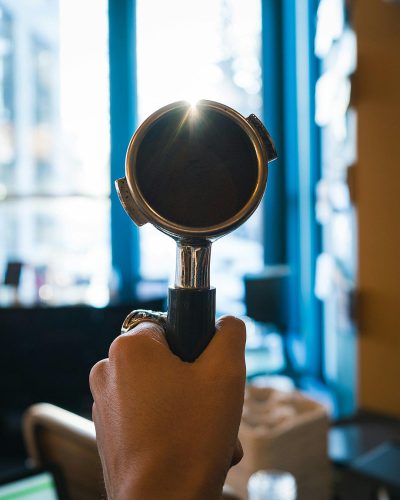 a hand holding an espresso portafilter in front of the camera, in modern cafe with blue window frames, sun shining through, shot on canon eos r5