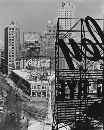 A black and white photograph of the CocaCola billboard on top, in downtown Los Angeles during its construction in early spring of 2035. The photo captures both cityscape and building details. In view is an enormous cocacola sign with neon letters on steel beams. A busy street below leads to buildings like "THE fluids Mayflower" or "the Genie ab subset". On one side there's another billboard for "CTaking you back", and on the other a large television screen showing news. It’s a sunny day.