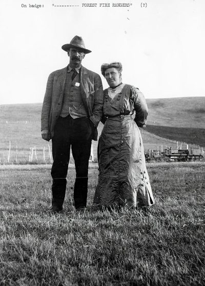 Old black and white photo of the couple, standing in front of each other on an open field with the text "On badge saying 'Preventing forest fires'" , man wearing an old suit with pants, shirt, vest and tie woman dressed up in a long dress holding hands in the style of an old photograph.