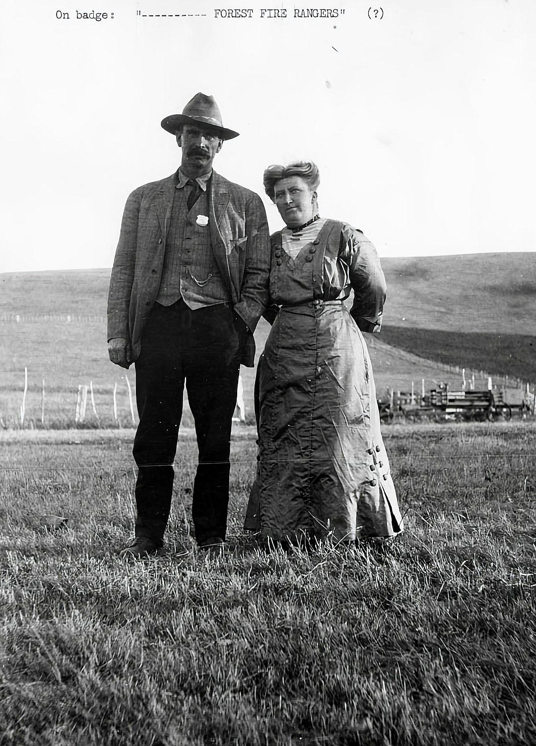 Old black and white photo of the couple, standing in front of each other on an open field with the text “On badge saying ‘Preventing forest fires'” , man wearing an old suit with pants, shirt, vest and tie woman dressed up in a long dress holding hands in the style of an old photograph.