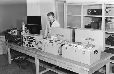 In the early days of radio, an engineer in white coat and black tie is working on some equipment placed at table with sign "YLE AAAllowed frantic movement between various energy and audio devices such as large studio and home shop sound peeled, and large boxes that have a small speaker and two oval wires sticking out from each side. The room also has shelves filled with other radio and technical tools. Black & White photo