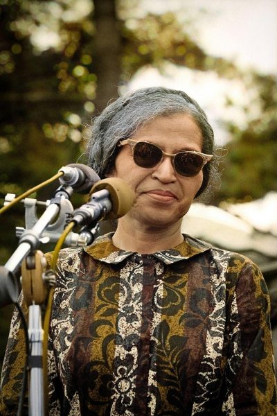 A photo of Rosa Parks, wearing sunglasses and an African print dress with matching shoes, giving her iconic speech at the bus boy's camp in tale as she stands on stage behind microphones for World day in Virginia, in the style of 20th century photography with an f/5.6 aperture setting to capture the entire scene in soft natural lighting.