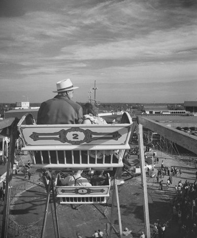 A man sitting on a high ride at a carnival, with a view of a crowd and racetrack in the background, in the style of a 1950s black and white photo by Alastair Scott, taken with a wide angle.