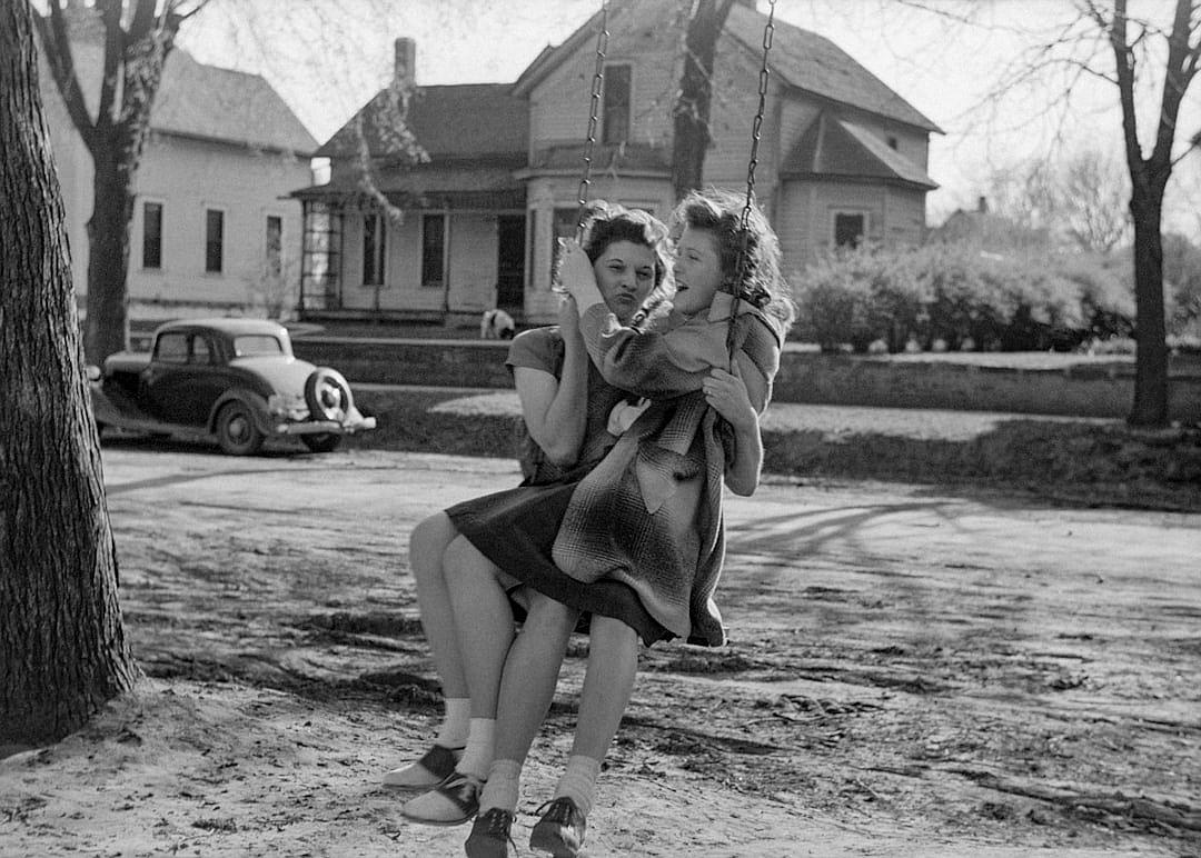 A black and white photograph of two women in the style of James Deborah Eoroiploch, one woman is sitting on another girl’s lap while she swinging from her arms outside their suburban home in an American town in early spring. The other lady has short curly hair wearing a dress with long sleeves that go down to just above knee length. There’s some grass around them, there are trees behind it, there’s a car parked next door, there’s yard space between house and street where you can see houses behind them. It was taken during midday sun, so shadows should be visible but not too harsh. The photo shows both