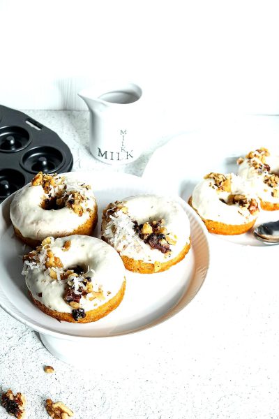 White background, side view of three white donuts with cream cheese glaze and walnut pieces on top. A black mug that says "MILK" is next to the plate. The kitchen counter has more plates, knives, a scale for weight control, and other cooking tools.