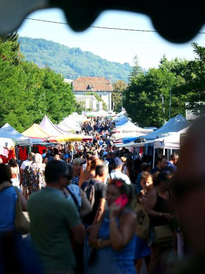 photo of an outdoor street market in the french countryside, crowds walking around, blue sky and green trees, white tents with pop up stalls selling art and goods, architecture in background, mountains visible in distance, shot from behind people's heads looking at camera,