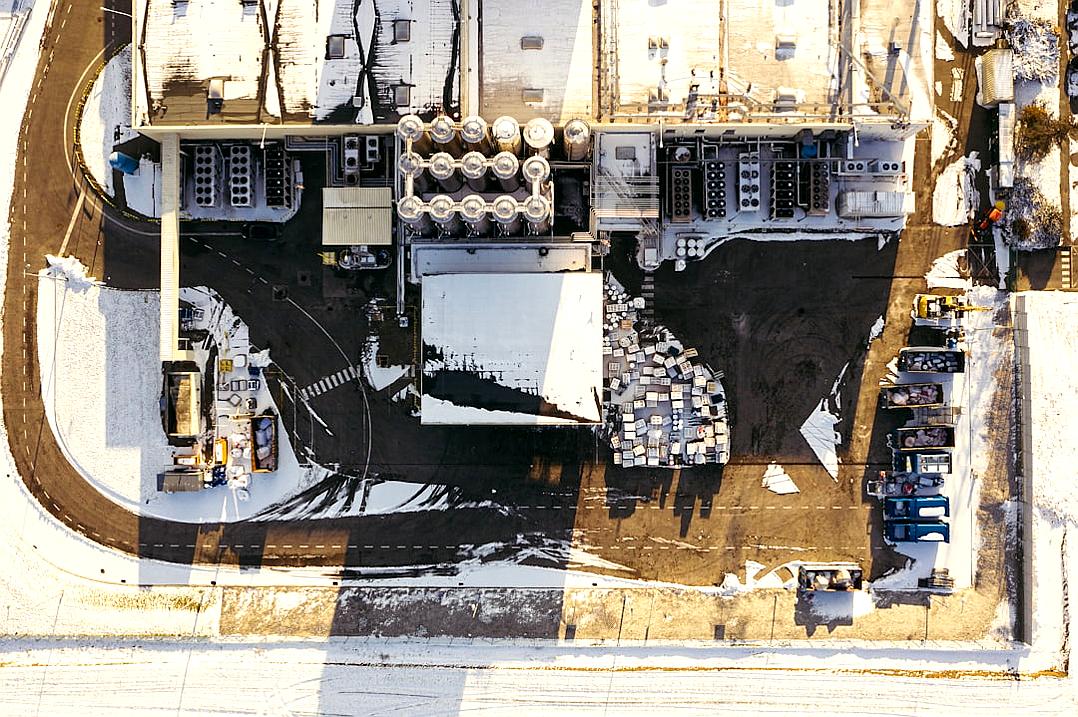 Aerial view of an industrial plant with many oil drums and workers in winter, on the ground is empty parking lot filled by snow, large truck loading cargo from one to another, white sky, realistic photography, detailed texture, professional color grading, soft shadows, no contrast, clean sharp focus, magazine photoshoot,