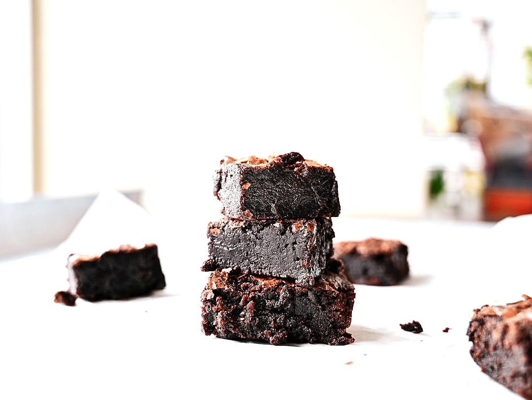 A photo of dark fudge brownies stacked on top of each other, placed in front of an empty white table with some cut up pieces scattered around the bottom left corner. The background is blurred and light grey, creating a soft focus effect.