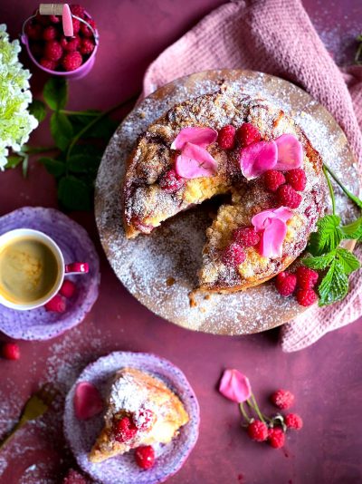 A beautiful photo of the delicious dish of pink crumb cake with vanilla, dusted in confectioner's sugar and fresh rose petals on top, placed beside it is an elegant purple plate containing coffee with cream and pink flowers, there are also some raspberries scattered around, the whole scene exudes romantic vibes, shot from above, taken in the style of canon eos r5.