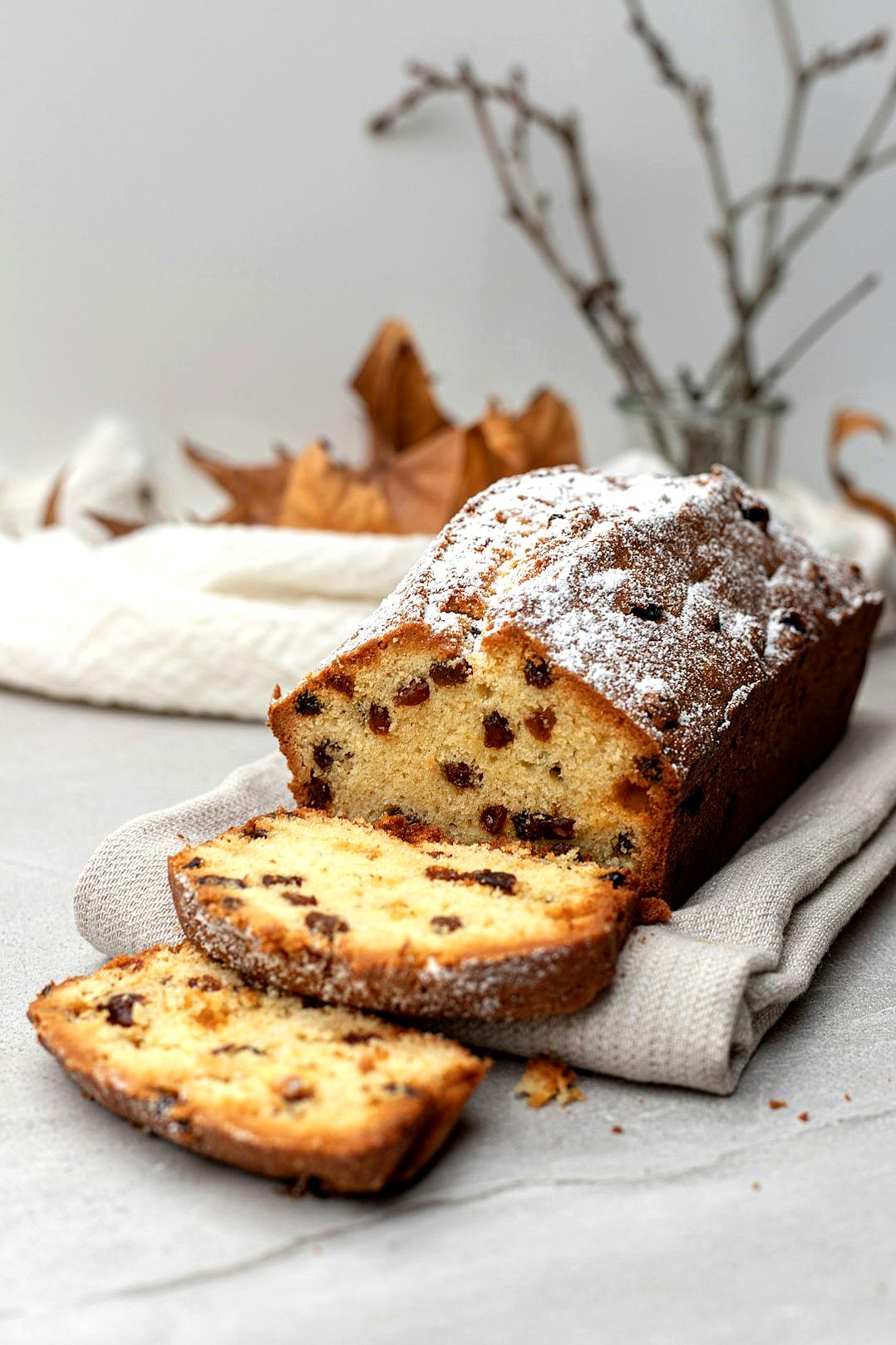 A photo of a plush loaf cake, dusted with flour and decorated with small dried fruits, is placed on a light gray tablecloth. A slice has been cut from it and lies next to it, with some chocolate chips visible in its texture. The background features dry branches, creating an atmosphere reminiscent of autumnal nature in the style of nature.