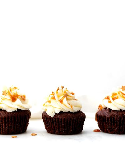 Chocolate cupcakes with white frosting and caramel drizzle, arranged in a line on the right side of an all-white background, a few pieces of coconut scattered around.