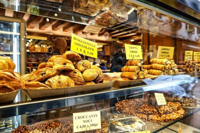 A display case in an Italian bakery displaying various pastries, including croissants shaped like wedges filled with chocolate and hazelnut cream. The sign on the glass says "Specialties by Kristina". In front of it is a stack of small transparent plastic bags full of nuts, behind which there's another pile containing large clear shades filled with coffee beans. There should be some people walking in the style of Kristina but they would not have to appear clearly visible or focused.