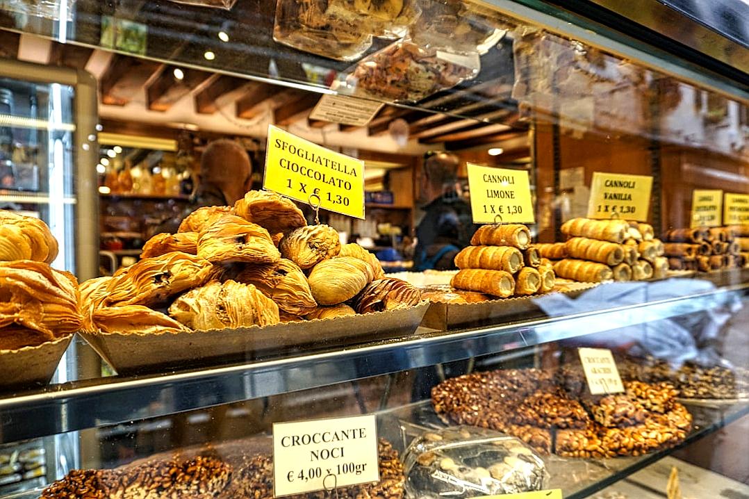 A display case in an Italian bakery displaying various pastries, including croissants shaped like wedges filled with chocolate and hazelnut cream. The sign on the glass says “Specialties by Kristina”. In front of it is a stack of small transparent plastic bags full of nuts, behind which there’s another pile containing large clear shades filled with coffee beans. There should be some people walking in the style of Kristina but they would not have to appear clearly visible or focused.