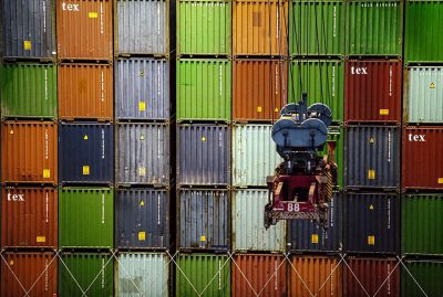 A person on top of a container is loading containers onto another, stacked with colorful shipping boxes labeled "'hex'". The background features numerous green and red metal shipping containers arranged in rows against an industrial setting. A forklift is visible near one of these large colored cargo containers. Captured using a Canon EOS5D Mark III camera with an EF lens at an f/8 aperture, ISO film speed set to F20, shutter time settings showing fast movement, and focal depth. The style captures the movement and industrial setting.