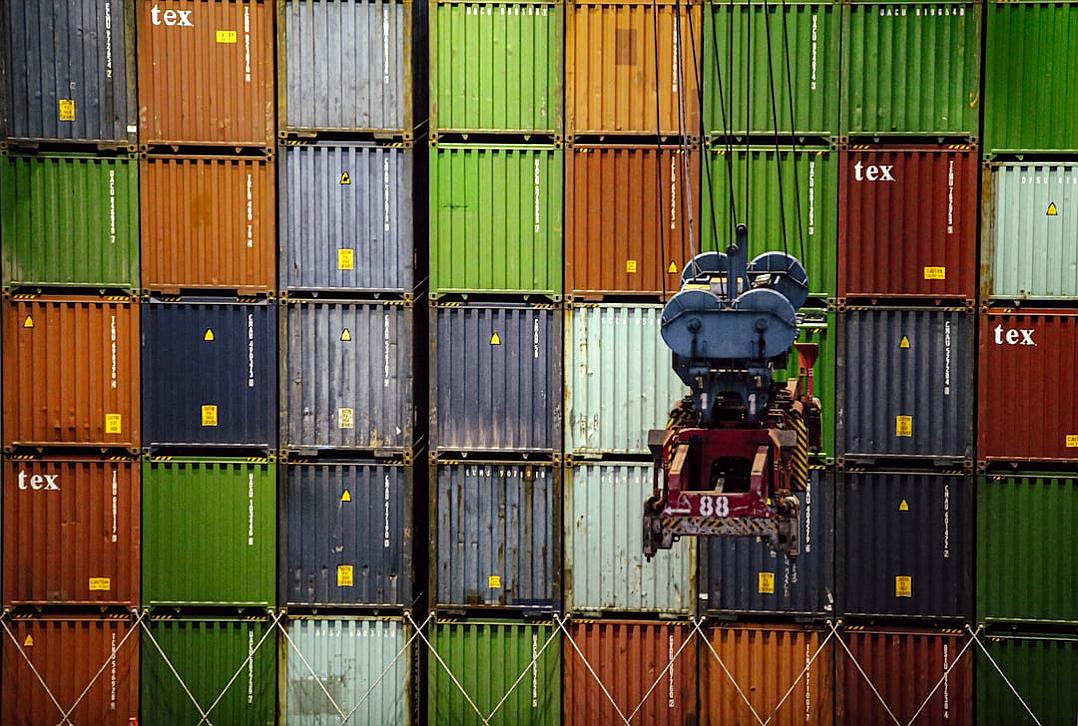 A person on top of a container is loading containers onto another, stacked with colorful shipping boxes labeled “‘hex'”. The background features numerous green and red metal shipping containers arranged in rows against an industrial setting. A forklift is visible near one of these large colored cargo containers. Captured using a Canon EOS5D Mark III camera with an EF lens at an f/8 aperture, ISO film speed set to F20, shutter time settings showing fast movement, and focal depth. The style captures the movement and industrial setting.