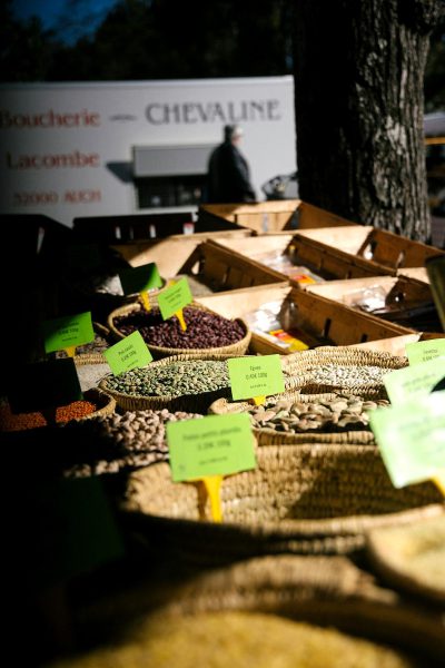 A photo of various beans and seeds in baskets at an outdoor market, with the words "Boulangerie de la chevaune" written on green signs next to them. A white truck is parked behind it. In the background there are some tree trunks. It’s evening light. Shot in the style of [Annie Leibovitz](https://goo.gl/search?artist%20Annie%20Leibovitz) with a Nikon D850 camera using Kodak Portra Pro 400 film. The colors are vibrant.