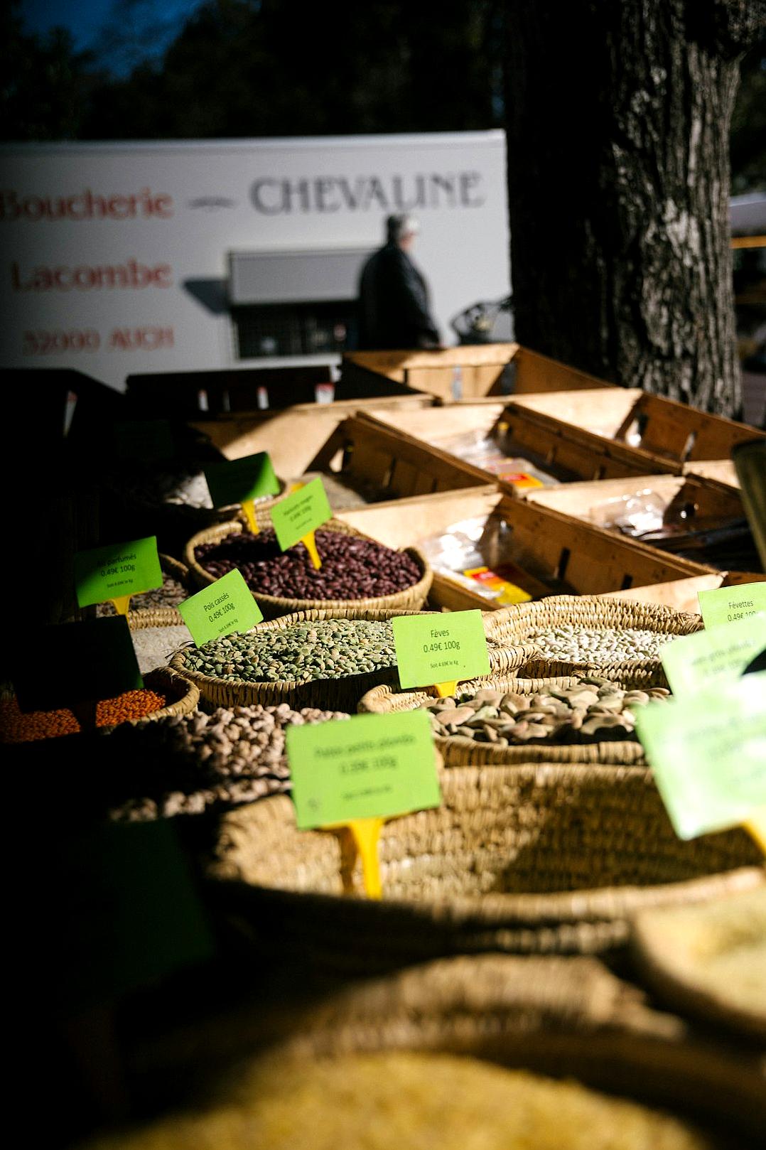 A photo of various beans and seeds in baskets at an outdoor market, with the words “Boulangerie de la chevaune” written on green signs next to them. A white truck is parked behind it. In the background there are some tree trunks. It’s evening light. Shot in the style of [Annie Leibovitz](https://goo.gl/search?artist%20Annie%20Leibovitz) with a Nikon D850 camera using Kodak Portra Pro 400 film. The colors are vibrant.