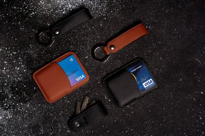 A topdown view of an array of leather keysters, featuring two different colors and styles of minimalistic leather card vertically positioned on the left side with their credit cards visible inside them, one in black color, another in brown color, while one slim blue minimalist wallet lies horizontally under it, all placed against a dark gray background, with soft lighting casting gentle shadows to highlight textures and details.