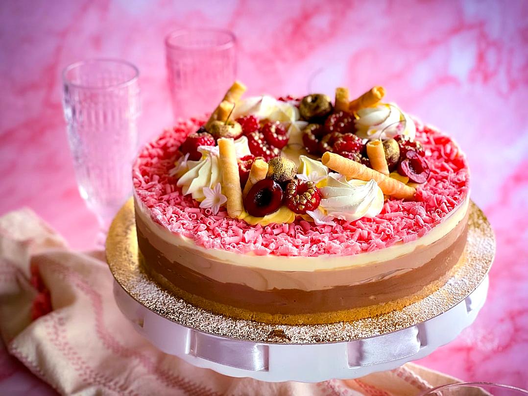 A delectable round cake adorned with creamy chocolate and cherry elements, topped with white cream decorations and fruit pieces on the pink textured surface. The setting includes two glasses filled with water in front of it, all set against an isolated pastel background.