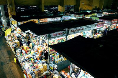 An aerial view of the interior of an old-fashioned food market from a top down perspective with a bird's eye view. The photorealistic style depicts a night time scene with large black roof and white signs and lights hanging from it. People are shopping for street foods such as scarves, fabrics, dried fruits, flowers, and dried plants. The colorful scene is in the style of National Geographic using a Nikon D850 DSLR camera with an aperture of f/4 and ISO set at 200.