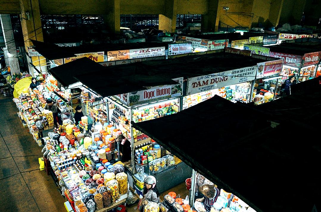 An aerial view of the interior of an old-fashioned food market from a top down perspective with a bird’s eye view. The photorealistic style depicts a night time scene with large black roof and white signs and lights hanging from it. People are shopping for street foods such as scarves, fabrics, dried fruits, flowers, and dried plants. The colorful scene is in the style of National Geographic using a Nikon D850 DSLR camera with an aperture of f/4 and ISO set at 200.