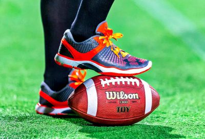 A closeup of an American football player's shoes standing on top of the ball, with the laces shifted to one side and adorned with bright orange laces, while he wears gray and blue sneakers featuring the Nike logo. The background is green grass at a roof garden stadium. Captured using a Canon EOS R5 camera with a macro lens, focusing on detail in the style of a realistic photography.