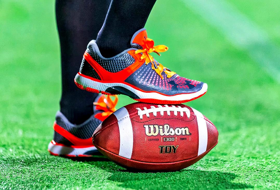 A closeup of an American football player’s shoes standing on top of the ball, with the laces shifted to one side and adorned with bright orange laces, while he wears gray and blue sneakers featuring the Nike logo. The background is green grass at a roof garden stadium. Captured using a Canon EOS R5 camera with a macro lens, focusing on detail in the style of a realistic photography.