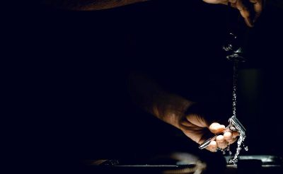 A hand turns on the tap, with water flowing down and dripping from it into an ashtray. The background is a dark black, highlighting only their hands in light. Captured using a Sony Alpha A7 III camera, focusing on capturing intricate details of fingers holding a cigarette holder and pouring liquid from a spout to a glass.