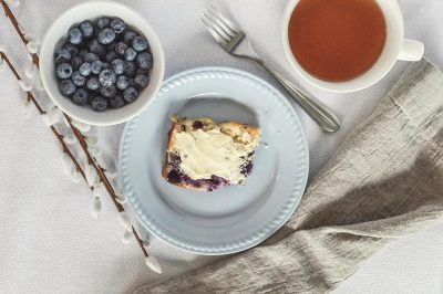 A photo of an elegant blueberry coffee cake, cut into small squares and served with butter on the side, placed in front of two willow branches on a white tablecloth, accompanied by fresh berries and a tea cup, captured from above using a Canon EOS camera in soft natural light and a minimalist style.