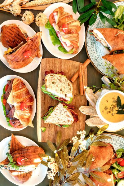 A spread of delicious and colorful sandwiches, including croissant BLTs with bacon on white plates. A wooden board is in the center holding one sandwich that has cheese melting down it. The background features various other dishes such as salad, soup, coffee cups, and fresh flowers, creating an inviting atmosphere for friends to share their laughter over good food.