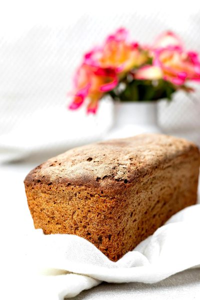 A photo of a whole loaf of homemade rye bread, sitting on a white kitchen towel with pink and orange flowers in the background in soft natural light.