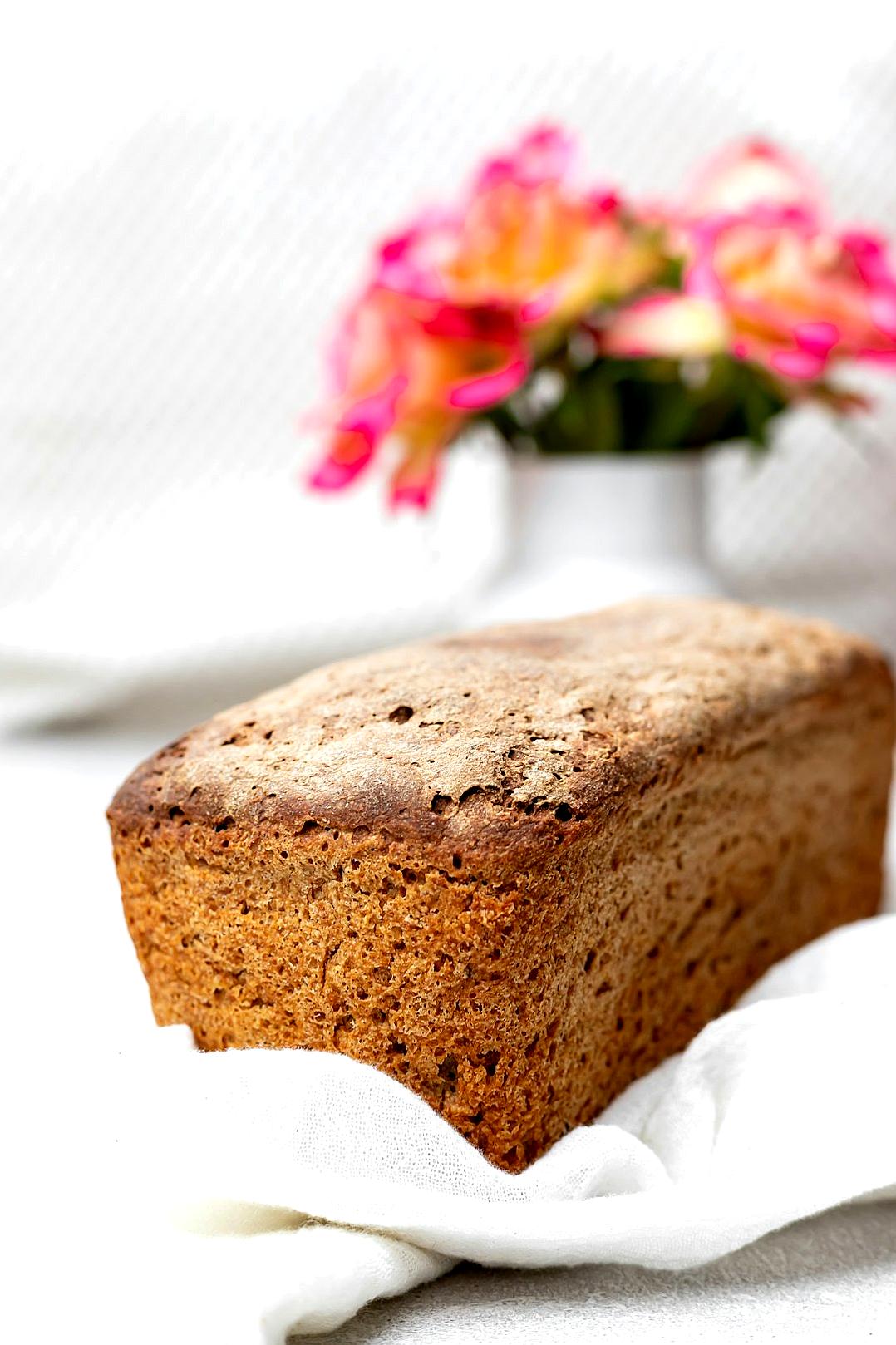 A photo of a whole loaf of homemade rye bread, sitting on a white kitchen towel with pink and orange flowers in the background in soft natural light.