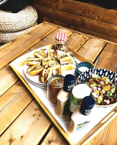 A wooden tray with white and blue checkered patterns, featuring various dishes such as hot dogs on buns, salad in bowls with spoons, jars of salt or pepper, and a bowl filled with cheese sauce., The background is an outdoor table with wood slats. The photo was taken from above at eye level, with natural lighting that highlights the colors of each dish and adds warmth to them.