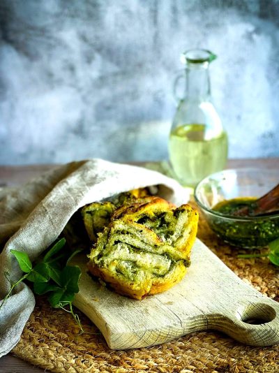 A professional food photograph of herby garlic sour cream stuffed bread, showcasing its vibrant green color and intricate texture against the soft backdrop of natural light. The setting includes an elegant wooden board with herbs scattered around it for decoration. A glass jar filled with mint leaves sits next to the bread in front of a textured white wall. This composition creates a warm and inviting atmosphere that highlights the unique combination of bread and garlic herb spread in the style of Larson's.