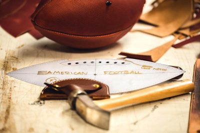 A leather football in the shape of an American football with "AMERICA excels" written on it lies next to one side and is being held in the style of hammers, tools for crafting armor made from natural materials. A design blueprint shows how these should look like they were used as helmets or gloves for playing American football.