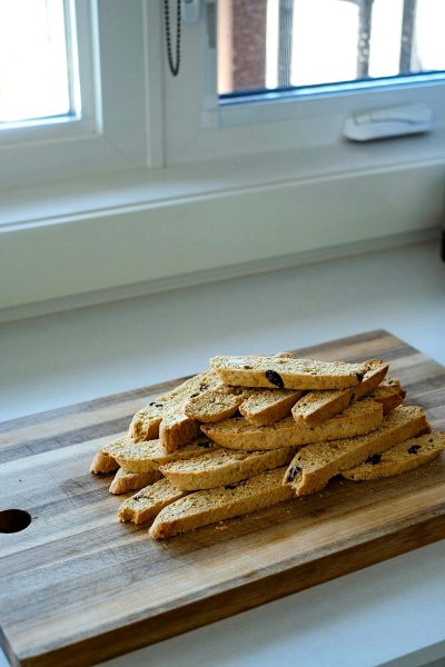 Biscotti on a wooden board on a kitchen counter with a window in the background, a white wall and floor, bright daylight.