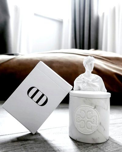 A white marble jar with the brand logo of "O & O" is placed on top, and inside it lies an embossed Roman statue figure in ivory color. The box stands next to the bed, and its lid rests against the headboard. It's a product photo for luxury candles, which can be seen through the transparent plastic case. There should not only be the text 'D' but also some small letters. This scene takes place in a bedroom with gray walls in the style of minimal editing.